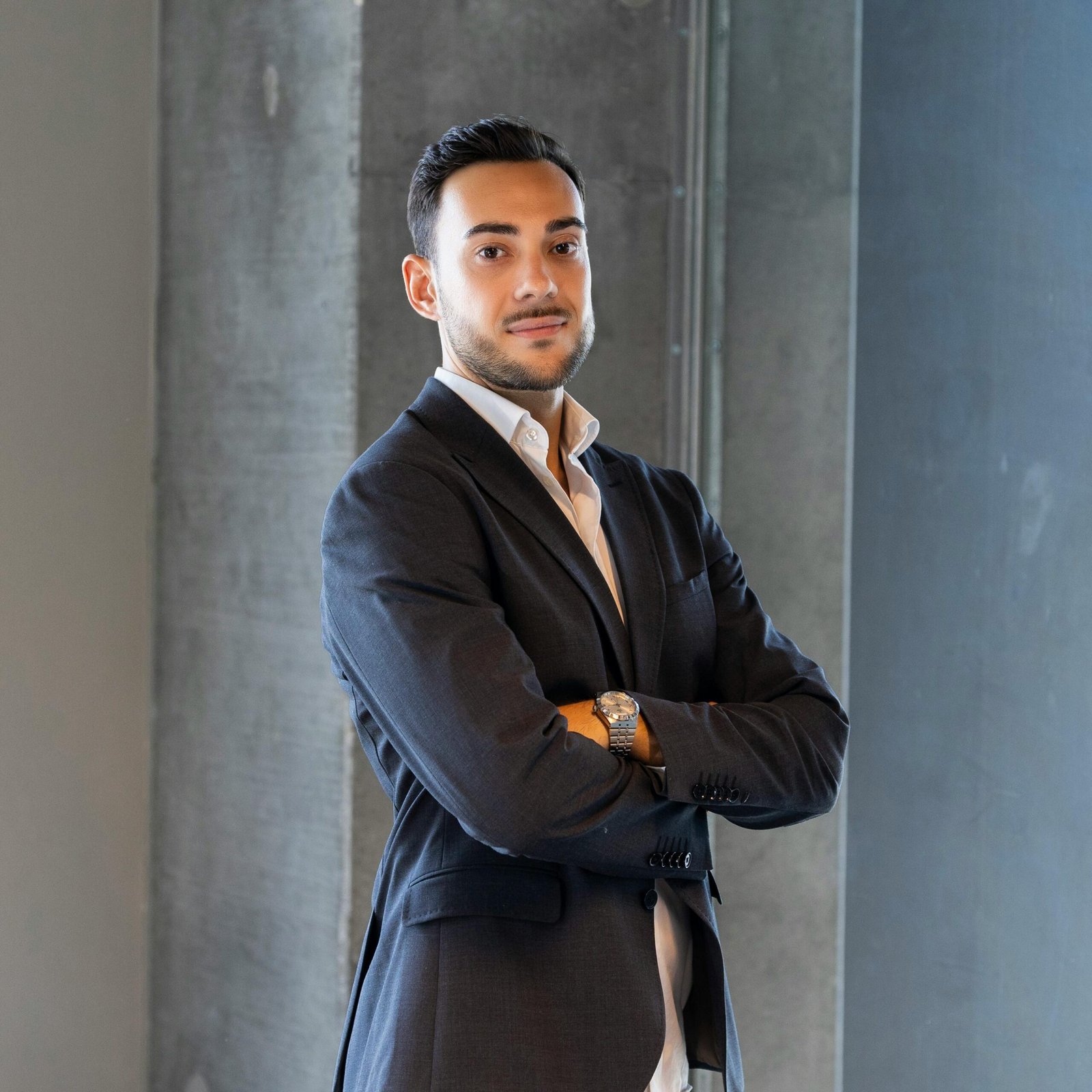 A poised businessman in formal attire standing confidently indoors with arms crossed.